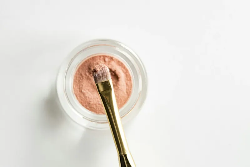 A pot of shimmering peach blush with a gold-handled makeup brush